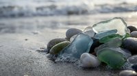 Smooth sea glass and pebbles scattered on sandy beach shore with gentle ocean waves in soft natural lighting creating peaceful coastal atmosphere.