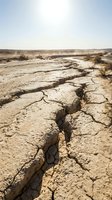 Arid landscape with deep cracks in parched earth, sparse vegetation, and water pooling in distance, depicting severe drought conditions and environmental degradation for climate change awareness.