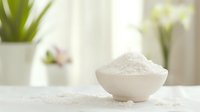 White sea salt crystals in ceramic bowl on light surface, soft natural window lighting creates minimalist spa atmosphere with blurred plant background.