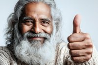 Senior Asian man with long gray beard and hair showing thumbs up gesture with warm genuine smile, wearing beige sweater against light background.