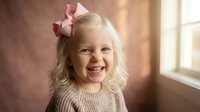 Happy blonde toddler girl with pink bow smiling at camera in cozy knit sweater by sunny window for family lifestyle photography and childhood memories.