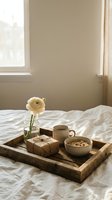Cozy breakfast in bed scene with wooden tray, coffee cup, granola bowl, wrapped gift and yellow ranunculus flower in glass vase on white bedding.