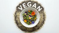 Chia seeds arranged in a circle spelling VEGAN around a white bowl filled with edible flowers and microgreens on white background.