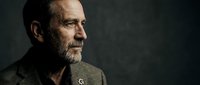 Mature man with beard and gray hair wearing olive blazer, looking thoughtfully to side against dark background, suitable for business, leadership, and professional portraits.