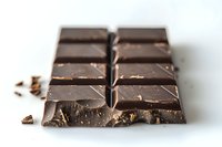 Dark chocolate bar broken into squares on white background with scattered crumbs, macro photography showing rich texture and glossy surface detail.