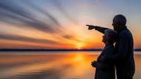 Father and son silhouettes pointing at sunset over calm lake water with dramatic orange and purple sky clouds reflecting on surface during golden hour.
