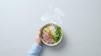Person holding steaming bowl of Asian noodle soup with meat, vegetables and lime garnish on white background, ready for food packaging and restaurant menu design.