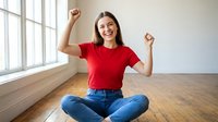 Young Hispanic woman in red shirt celebrating success with raised fists while sitting cross-legged on wooden floor in bright modern room with large windows.