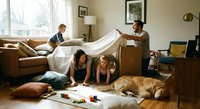 Family building blanket fort in cozy living room with children playing underneath while parents help create magical indoor hideaway for quality time.