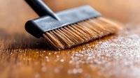 Soft bristle brush sweeping white powder or flour on wooden surface in macro photography, creating artistic texture and movement with shallow depth of field.