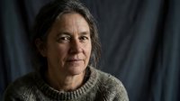 Middle-aged woman with long dark hair wearing a knit sweater, looking thoughtfully at camera against dark draped background for portrait use.