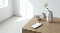 Minimalist wooden desk with smartphone, ceramic bowls and white vase in bright modern interior with natural sunlight streaming through window.