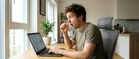 Man with curly brown hair, Caucasian ethnicity, wearing olive green t-shirt, sitting at desk with laptop, hand near face in thoughtful pose, working from home office with natural light from windows.