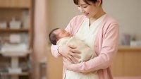 Young Asian mother in pink cardigan gently holds sleeping newborn baby, warm home interior background, parenthood and family care concept.