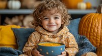 Cheerful child with curly hair holding orange ceramic mug with hot drink, wearing cozy polka dot hoodie, sitting on sofa with autumn pumpkin decor background.