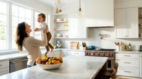 Happy mother lifting toddler in bright modern kitchen with white cabinets and marble countertops during morning routine.