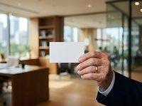 Mature male professional holding blank white business card in modern office environment with wooden furniture and glass partitions.
