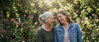 Senior woman with gray hair and adult daughter with blonde hair smiling together in blooming garden with pink flowers and green foliage, representing family bonding and generational connection.