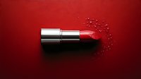 Red lipstick with scattered sparkles and glitter particles on dark red background. Luxury cosmetics product photography for beauty and fashion advertising.