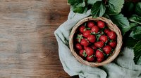 Fresh ripe strawberries in wicker basket on rustic wooden background with green leaves and linen cloth, top view food photography.