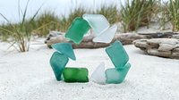 Recycling symbol made from sea glass and frosted glass pieces arranged on sandy beach with driftwood and beach grass, representing environmental conservation and sustainable living practices.