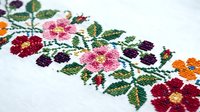 Colorful floral cross stitch embroidery with pink purple orange flowers and green leaves on white fabric for traditional handicraft projects.