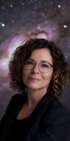 Caucasian woman with curly brown hair wearing glasses and black sparkly outfit smiles warmly against dreamy bokeh background in pink and purple tones.