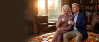 Happy senior Caucasian couple enjoying coffee together in cozy living room with warm sunlight streaming through window creating intimate domestic moment.
