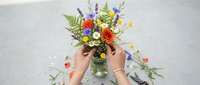 Hands holding vibrant mixed flower bouquet in glass vase with colorful blooms and greenery on white surface for floral design and decoration projects.