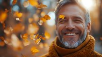Mature Caucasian man with gray beard smiling outdoors while golden autumn leaves fall around him, wearing brown scarf, captured in warm sunset lighting.