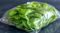 Fresh green basil leaves in transparent plastic bag on dark background, macro food photography showing texture and freshness of aromatic herbs.