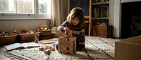 Young blonde girl playing with wooden building blocks on living room floor, focused on stacking and construction play in cozy home interior with natural light.