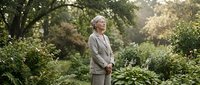 Senior Caucasian woman in beige cardigan standing peacefully in lush garden surrounded by green foliage and plants during golden hour sunlight.