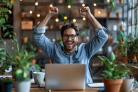 Excited young professional celebrating success at laptop in cozy cafe setting with plants and warm lighting. Perfect for business, entrepreneurship, and remote work concepts.