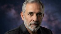 Middle-aged man with gray hair and beard wearing dark clothing against blurred purple background. Suitable for corporate portraits, professional headshots, business profiles.