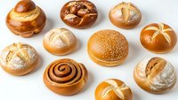 Assorted fresh baked bread rolls and pastries including sesame burger buns, cinnamon swirls, pretzels and artisan loaves on white background.