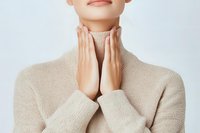 Young Caucasian woman in beige turtleneck sweater touching her neck with both hands, showing skincare and wellness concept against light background.