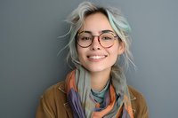 Young woman with platinum blonde hair and round glasses smiling warmly at camera, wearing colorful scarf and brown jacket against gray background. Natural daylight portrait.
