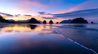 Dramatic sunset over ocean beach with rocky sea stacks and colorful sky reflecting on wet sand creating mirror effect during golden hour twilight.