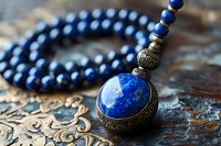 Vintage bronze pendant with deep blue lapis lazuli stone and matching beaded necklace on ornate metallic surface, showcasing intricate antique jewelry design and craftsmanship.