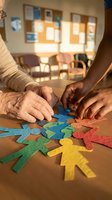 Senior adult and young adult with dark skin tones assembling colorful paper figures together at wooden table in bright classroom setting, promoting intergenerational bonding and cooperative learning activities.