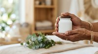 Close-up of hands holding blank white jar with fresh herbs on wooden table in bright kitchen, suitable for product packaging mockups and wellness branding.