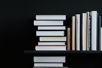 Stack of books arranged on dark shelf against black background, minimalist composition with white and neutral colored book spines creating visual contrast.