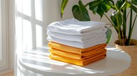 Fresh white and orange folded bed linens stacked neatly on round table near window with monstera plant, bright natural daylight creating cozy home atmosphere.