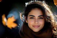 Young Middle Eastern woman with natural makeup smiling warmly while wearing brown scarf, autumn maple leaves blurred in background creating seasonal outdoor portrait.