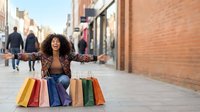 Young African American woman sitting on city street surrounded by colorful shopping bags, arms wide open, laughing joyfully on urban shopping street.