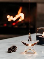 Glass star ornament glowing against blurred fireplace background with pinecone on marble surface, creating cozy winter holiday atmosphere.