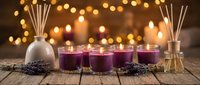 Purple lavender scented candles and reed diffusers arranged on rustic wooden table with warm bokeh lights creating cozy spa atmosphere for relaxation.