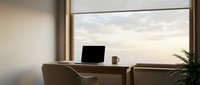 Laptop and coffee cup on wooden desk by large window with sunset sky view, workspace setup for remote work and productivity.