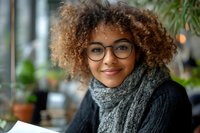 Young African American woman with curly hair and glasses smiling warmly in cozy cafe setting with knitted scarf for lifestyle and portrait photography.